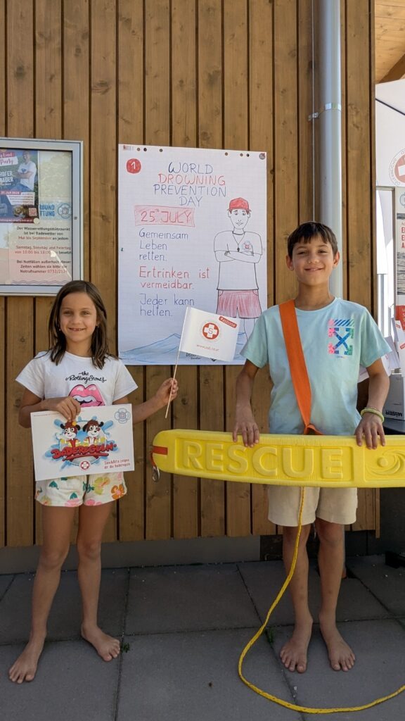 Zwei Kinder stehen vor einem Holzwand-Plakat zum World Drowning Prevention Day und halten eine gelbe Rettungsboje.